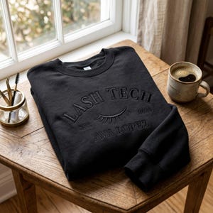 May include: Black crewneck sweatshirt with the text "LASH TECH" above an eyelash graphic, "EST. 2024 AVA LOPEZ" below. The sweatshirt is folded on a wooden table, next to a cup of coffee and a small glass jar with tools.