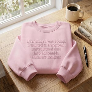 Puede incluir: Una sudadera rosa claro con el texto "Ever since I was young, I wanted to transform unstructured data into actionable business insights." Una taza de café, libros y un bolígrafo sobre una mesa de madera.