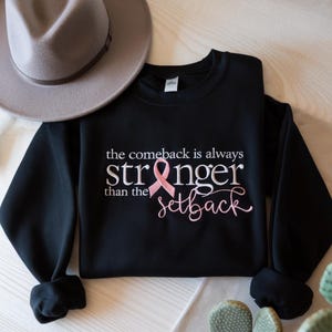 Könnte beinhalten: Schwarzes Sweatshirt mit dem Text "the comeback is always stronger than the setback" in Weiß und Rosa. Ein rosa Band ist in das Design integriert. Ein grauer Hut ist im Hintergrund.
