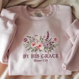 Pode incluir: Camisola rosa claro com design floral bordado em rosa, roxo e verde. O texto "BY HIS GRACE Romans 3:24" é bordado abaixo das flores. A camisola tem mangas compridas e gola redonda.