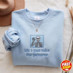 Könnte beinhalten: Ein hellblaues Sweatshirt mit Rundhalsausschnitt und einem gestickten quadratischen Aufnäher, der ein winkendes Skelett zeigt. Unter dem Aufnäher befindet sich der in Blau gestickte Text "idk i just take the pictures". Das Kleidungsstück hat gerippte Bündchen.