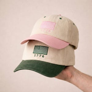 May include: Two baseball caps stacked vertically. The top cap is beige with a pink brim and features an embroidered pink American flag and the text 1776. The bottom cap is beige with a dark green brim, featuring an embroidered green American flag and 1776.