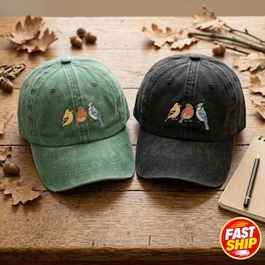 Puede incluir: Dos gorras de béisbol, una en verde salvia y otra en gris carbón, cada una con un diseño bordado de tres pájaros pequeños en amarillo, naranja y azul. Las gorras tienen una corona estructurada, una visera curva y una construcción clásica.