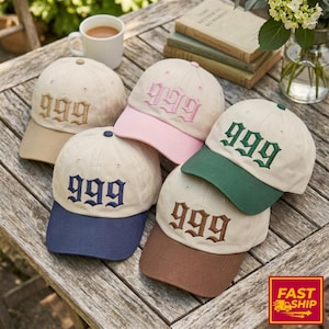 May include: A collection of beige baseball caps with the number "999" embroidered on the front in various colors, including brown, pink, green, and blue. Each cap has a different colored brim. The caps are arranged on a wooden surface.