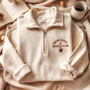 Op de afbeelding: Crèmekleurige sweatshirt met een kraag met kwartrits en de geborduurde zin "MAYBE COFFEE IS ADDICTED TO ME." Een kleine afbeelding van een koffiekopje staat onder de tekst. De sweatshirt is opgevouwen op een zacht, neutraal getint oppervlak, met een koffiemok en een open boek op de achtergrond.