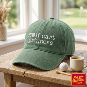 以下が含まれることがあります： 白い文字で「golf cart princess」と書かれた緑色の野球帽。ゴルフボール、木製のティー、小さなコーヒーカップが帽子の隣にあります。帽子は木製の表面にあります。