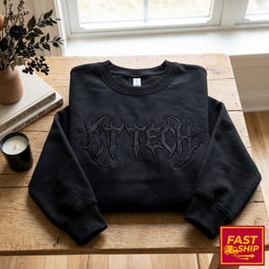 May include: Black crew neck sweatshirt featuring the embroidered text "DT TECH" in a gothic font. The sweatshirt is displayed on a wooden table, with a vase of flowers, books, and a candle in the background. The sweatshirt has ribbed cuffs and hem.