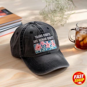 May include: A charcoal grey baseball cap featuring the embroidered white text "TRANS RIGHTS ARE HUMAN RIGHTS" above a rectangular patch displaying pink flowers with green leaves on a light blue background, with visible stitching on the brim.