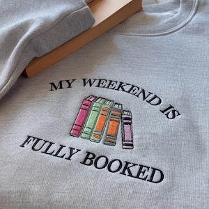 以下が含まれることがあります： 「MY WEEKEND IS FULLY BOOKED」の刺繍が施されたライトグレーのスウェットシャツ。カラフルな本が並んだデザインです。スウェットシャツは本の上に折りたたまれています。