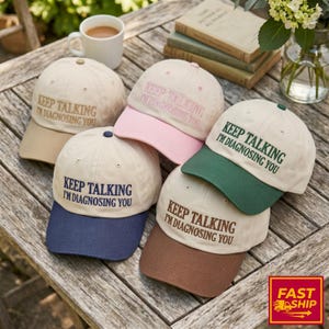 May include: Five baseball caps with off-white crowns and contrasting brims in tan, pink, dark blue, dark green, and brown. Each cap features the embroidered text "KEEP TALKING I'M DIAGNOSING YOU" in a color matching its respective brim.