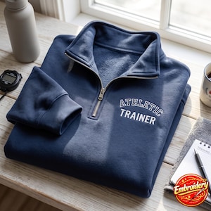 May include: A navy blue quarter-zip pullover sweatshirt featuring a high collar with a silver metal zipper. The chest area displays the white embroidered text "ATHLETIC TRAINER" in a bold, arched collegiate-style font on the right side.