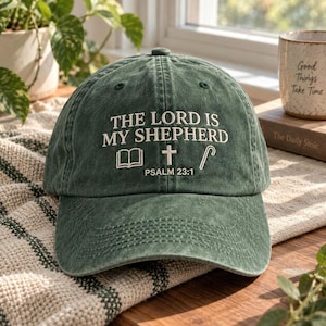 Puede incluir: Una gorra de béisbol verde bosque con acabado desgastado. El frente presenta un bordado blanco con el texto THE LORD IS MY SHEPHERD, seguido de iconos de un libro abierto, una cruz y un cayado, con el texto PSALM 23:1 debajo.