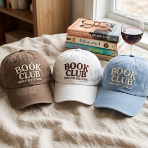 May include: Three baseball caps in brown, white, and blue. Each cap has the words "BOOK CLUB" embroidered in a contrasting color, with "HERE FOR THE WINE" below. The caps are on a neutral-toned surface, with books and a wine glass in the background.