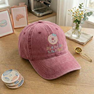 Könnte beinhalten: Staubrosa Baseballkappe mit gebogenem Schirm. Die Kappe hat einen gestickten Donut mit blauer Schleife und dem Text "It's A Bad Day To Be A Donut". Die Kappe liegt auf einer Holzoberfläche.
