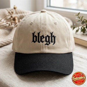 May include: A two-tone baseball cap featuring a beige crown and a black brim. The front of the crown displays the word blegh embroidered in black thread using a gothic-style font. The cap includes a top button and stitched eyelets on the panels.