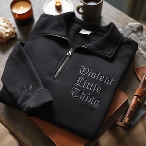 K&ouml;nnte beinhalten: Schwarzes Sweatshirt mit Viertelrei&szlig;verschluss und dem gestickten Text "Violent Little Thing" in gotischer Schrift. Das Sweatshirt hat einen silbernen Rei&szlig;verschluss und einen kleinen gestickten Drachen am &Auml;rmel. Es liegt auf einem braunen Leder-Notizbuch.