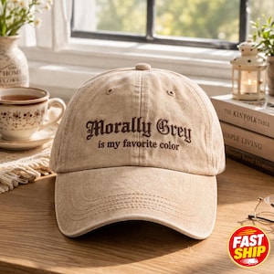 Może przedstawiać: Beżowa czapka z daszkiem o spranym, postarzanym wyglądzie. Z przodu znajduje się ciemnobrązowy haft z napisem Morally Grey is my favorite color wykonanym ozdobną czcionką szeryfową. Czapka posiada zakrzywiony daszek oraz guzik na górze.