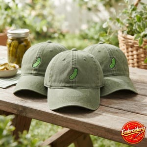 May include: Three olive green baseball caps made of cotton, each featuring a small, embroidered green pickle design centred on the front panel. The caps have a structured crown, a curved brim, and a classic six-panel construction with eyelets.