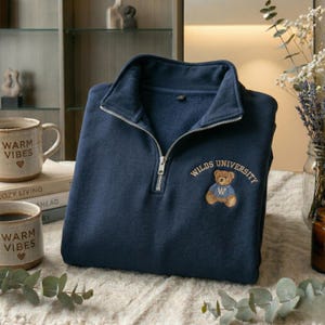 May include: Navy blue sweatshirt with a quarter-zip closure and a stand-up collar. The sweatshirt features an embroidered teddy bear graphic with the text "WILDS UNIVERSITY". Two mugs with the text "WARM VIBES" are visible.