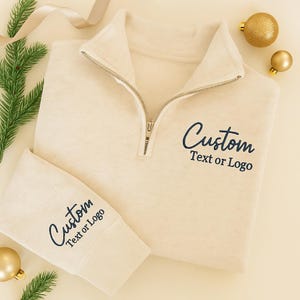 May include: Cream-colored quarter-zip sweatshirt with the text "Custom Text or Logo" in navy blue. The sweatshirt has a ribbed cuff and collar. The image is styled with gold ornaments and greenery.