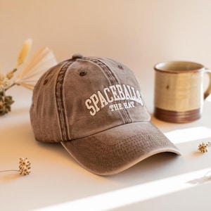 以下が含まれることがあります： 白文字で「SPACEBALLS THE HAT」と書かれた茶色の野球帽。白い表面に置かれ、茶色のマグカップの隣にあります。