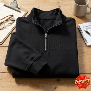 Puede incluir: Una sudadera negra con cremallera de un cuarto y la palabra RADIOLOGY bordada en hilo negro a juego en el pecho izquierdo. La prenda cuenta con un tirador de cremallera de metal plateado y mangas largas con puños acanalados.