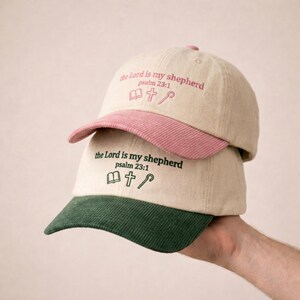 May include: Two baseball caps stacked vertically. The top cap is beige with a pink corduroy brim and pink embroidery reading "the Lord is my shepherd psalm 23:1" with icons of a book, cross, and staff. The bottom cap is beige with a dark green corduroy brim and dark green embroidery reading "the Lord is my shepherd psalm 23:1" with icons of a book, cross, and staff.