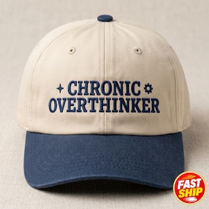 May include: A beige baseball cap with a dark blue brim and a dark blue button on top. The front features dark blue embroidered text that reads CHRONIC OVERTHINKER, flanked by a small four-pointed star symbol on the left and a gear icon on the right.