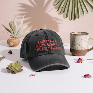 May include: A dark gray baseball cap with the text "I SUPPORT A MAN'S RIGHT TO SHUT THE FUCK UP" in red. The hat is made of a soft material and has a curved brim. A ceramic mug and decorative plants are in the background.
