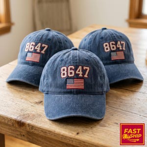 Könnte beinhalten: Drei blaue Jeans-Baseballkappen. Jede Kappe hat die Zahl "8647" in Rot und Weiß sowie eine kleine amerikanische Flagge. Die Kappen liegen auf einer Holzoberfläche.