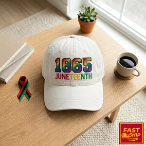 May include: A cream-coloured baseball cap with the text "1865 JUNETEENTH" in rainbow colours. A small ribbon in the colours of the Pan-African flag is next to the cap. A cup of coffee and a small plant are also visible.