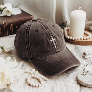 Puede incluir: Gorra de béisbol gris oscuro con una cruz blanca bordada en la parte delantera. La gorra tiene una visera curva y un diseño clásico de seis paneles. El sombrero está hecho de una tela suave y desgastada, y es un accesorio religioso.