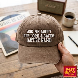 May include: A brown baseball cap with a distressed fabric finish featuring white embroidered text that reads ASK ME ABOUT OUR LORD & SAVIOR [ARTIST NAME] centered on the front panel, with a curved brim and a structured six-panel design.