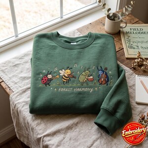 May include: A forest green crewneck sweatshirt featuring an embroidered graphic of four insects playing musical instruments, including a ladybug with a trumpet, a bee with a violin, a beetle with a drum, and a stag beetle with a cello, with the text Forest Harmony.