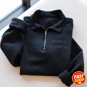 May include: A black long-sleeve quarter-zip sweatshirt featuring a fold-over collar and a metallic zipper. The text "Women in Engineering" is embroidered in a matching black thread on the right side of the chest in a gothic-style font.