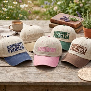 May include: Five beige baseball caps with embroidered text. The caps have different coloured brims, including blue, pink, green, and brown. The text on the caps reads "SOMEBODY" and "SOMEBODY'S PROBLEM".
