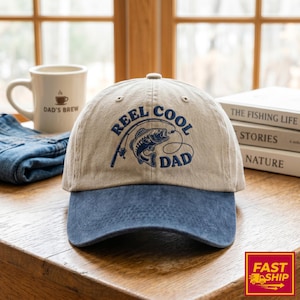 Op de afbeelding: Een beige en blauwe baseballcap met de tekst "REEL COOL DAD" en een visafbeelding. De cap heeft een gebogen klep en is gemaakt van een zachte stof. Een witte mok met "DAD'S BREW" staat op de achtergrond.