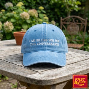 Op de afbeelding: Lichtblauwe baseballcap met de witte tekst "I AM BILLING YOU FOR THIS CONVERSATION". De pet ligt op een houten tafel. De achtergrond toont groen en een decoratieve stoel.