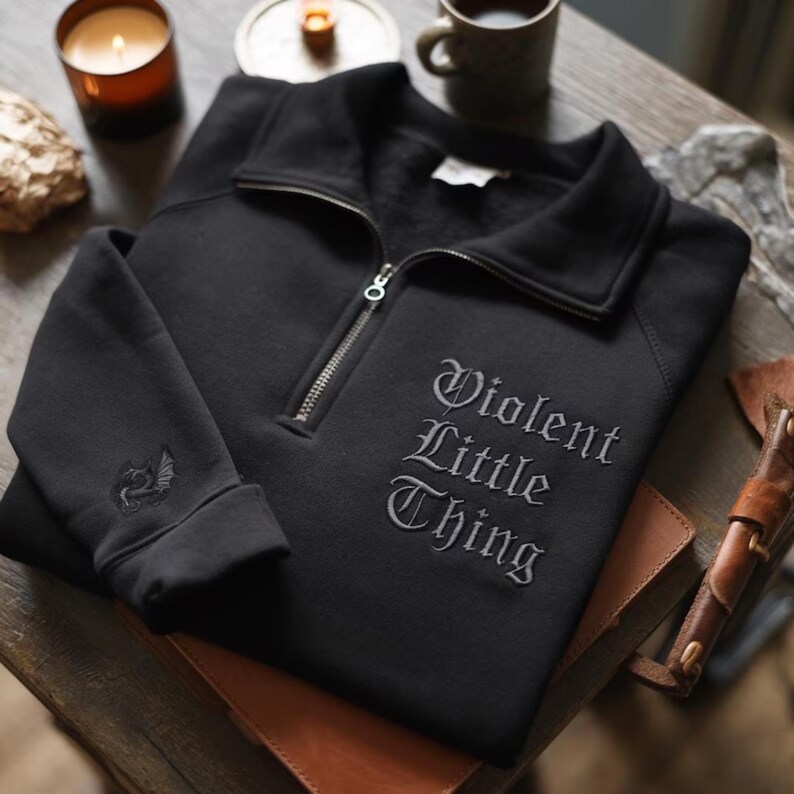 K&ouml;nnte beinhalten: Schwarzes Sweatshirt mit Viertelrei&szlig;verschluss und dem gestickten Text "Violent Little Thing" in gotischer Schrift. Das Sweatshirt hat einen silbernen Rei&szlig;verschluss und einen kleinen gestickten Drachen am &Auml;rmel. Es liegt auf einem Leder-Notizbuch.