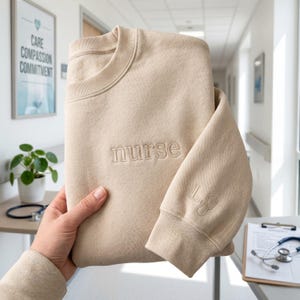 Puede incluir: Sudadera beige con la palabra "nurse" bordada en el pecho. La manga presenta un estetoscopio y las iniciales "L" y "J". La sudadera se sostiene frente a un pasillo.