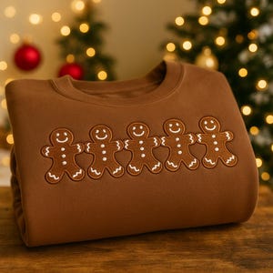 Może przedstawiać: Brązowa bluza z haftowanym wzorem pięciu ludzików z piernika trzymających się za ręce. Piernikowe ludziki mają białe detale z lukru i uśmiechnięte twarze. Bluza wykonana jest z miękkiego materiału.