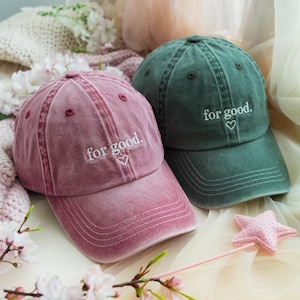 May include: Two baseball caps, one in dusty rose and one in dark green. Each cap features the embroidered phrase "for good." in white, with a small heart. The caps are made of a soft, textured fabric.
