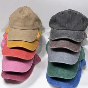 May include: Two vertical stacks of baseball caps in various colors. The left stack features tan, mustard yellow, red, pink, and maroon caps. The right stack includes charcoal, brown, light gray, dark green, and blue caps. All caps have a curved brim.