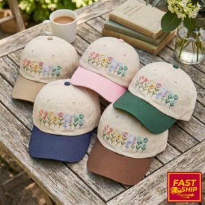Op de afbeelding: Een verzameling beige baseballpetten met geborduurde bloemmotieven. Elke pet heeft een klep in een andere kleur, waaronder beige, roze, groen, bruin en marineblauw. De petten liggen op een houten oppervlak.