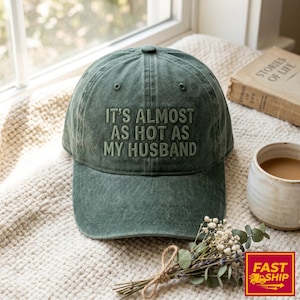 May include: A green baseball cap with the embroidered text "IT'S ALMOST AS HOT AS MY HUSBAND." The hat is made of a soft, worn material. The cap is sitting on a cream-coloured blanket.