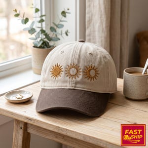 May include: A beige baseball cap with a brown peak and top button. The cap features embroidered sun and moon designs in yellow and brown. The cap is displayed on a wooden table.