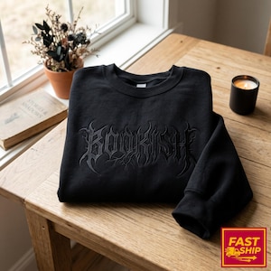 Könnte beinhalten: Schwarzes Sweatshirt mit Rundhalsausschnitt und dem Wort "BOOKISH" in gotischer Schrift gestickt. Das Sweatshirt liegt gefaltet auf einem Holztisch, im Hintergrund eine brennende Kerze und eine kleine Topfpflanze. Der Text "FAST SHIP" befindet sich unten rechts.