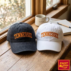 May include: Two baseball caps, one black and one white, sit side-by-side on a wooden surface. Both caps feature the word "TENNESSEE" embroidered in orange block letters. A coffee mug with "VOLS" printed on it and a notebook are in the background.