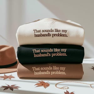May include: A stack of three sweatshirts in cream, black, and brown. Each sweatshirt has the embroidered phrase "That sounds like my husband's problem." A brown hat and autumn leaves are in the background.