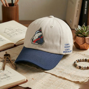 May include: A beige baseball cap with a navy blue brim. The cap features a patch with a graphic design and the text "COPY HEART COPY BRACELET" on the side. A bracelet and open book are in the background.